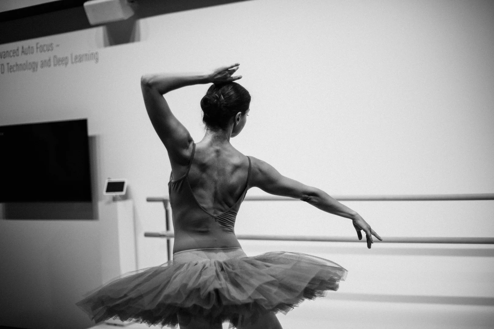 A dancer in a tutu caught mid-gesture at the studio barre in stark black and white, illustrative of Mats Ek's practice of taking apart classical ballet vocabulary and rebuilding it from within