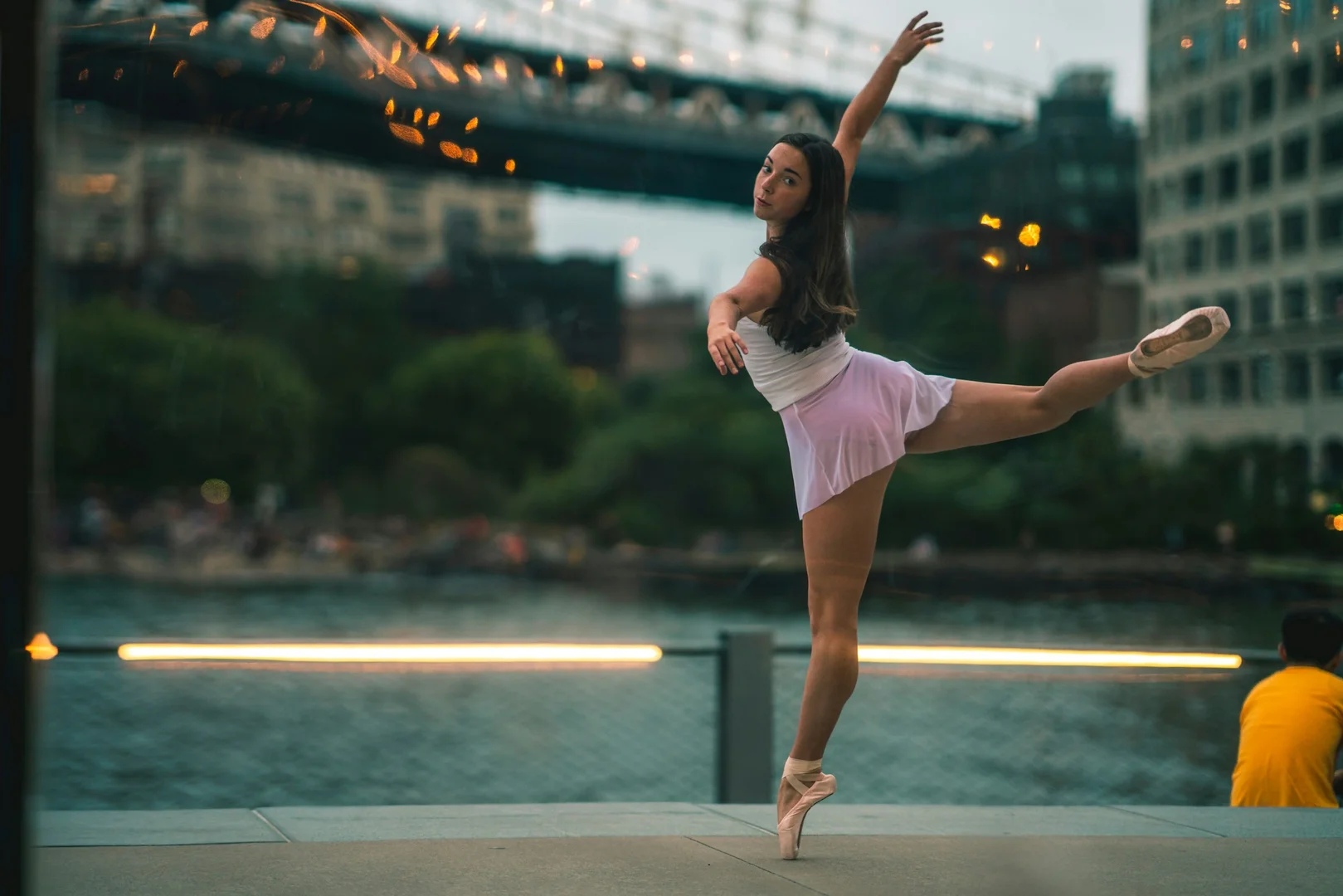 A solo dancer suspended in an extended arabesque against a city waterfront, illustrative of the theatrical, image-driven spectacle at the center of Alexander Ekman's work